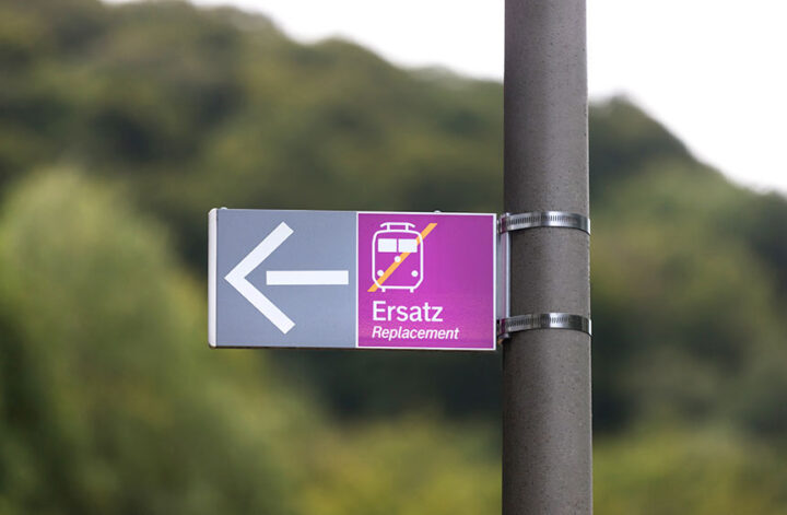 Foto zum Blogartikel "Schienenersatzverkehr Berlin–Hamburg: Orientierung, die Zeit spart": Ein Hinweisschild am Mast zeigt nach links und kündigt „Ersatz / Replacement“ an: lila Feld mit durchgestrichenem Zug-Piktogramm – Hinweis auf Schienenersatzverkehr. Unscharfer, grüner Hintergrund.